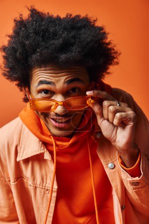 Homme afro-américain à la mode avec des cheveux afro bouclés et des lunettes de soleil posant expressivement dans une chemise orange.