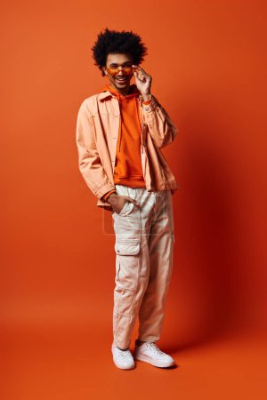 Foto de Un joven afroamericano de moda y emocional se levanta con confianza contra un fondo naranja brillante con atuendos y gafas de sol de moda.. - Imagen libre de derechos
