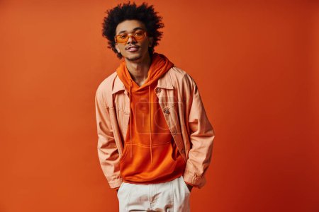 Ein lockiger afroamerikanischer Mann, der Stil und Emotion ausstrahlt, in trendigem orangefarbenem Hemd, weißen Shorts und Sonnenbrille auf orangefarbenem Hintergrund.