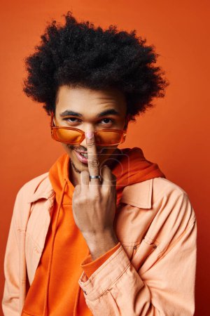 Téléchargez les photos : Un jeune Afro-Américain aux cheveux bouclés revêtu d'une chemise orange, d'une veste et de lunettes de soleil sur fond orange, mettant en valeur un style vibrant et éclectique. - en image libre de droit