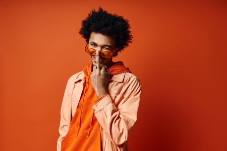 Foto de Joven afroamericano con estilo, pelo rizado, camisa naranja, manos en la cara, expresando emoción, fondo de moda. - Imagen libre de derechos