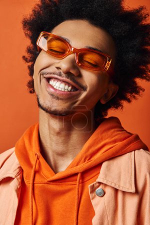 Stylish young African American man with curly hair, trendy attire, and sunglasses, smiling on orange background.