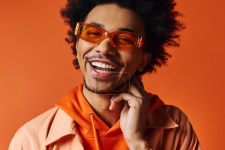 Téléchargez les photos : Un jeune homme afro-américain aux cheveux bouclés portant des vêtements et des lunettes de soleil à la mode, montrant un sourire lumineux à la caméra sur un fond orange. - en image libre de droit