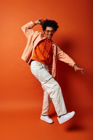A trendy African American man with curly hair and sunglasses, dressed in an orange shirt and white pants, poses against an orange background.