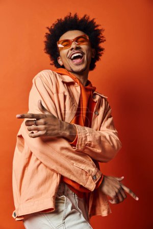 Stylish African American man with curly hair wearing sunglasses, showcasing emotions on an orange background.