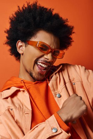 Homme afro-américain élégant aux cheveux afro en chemise orange et lunettes de soleil, respirant la fraîcheur sur fond orange.