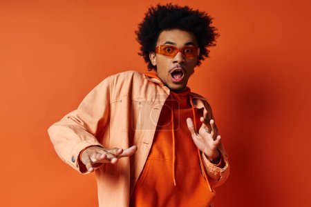 Ein stilvoller junger afroamerikanischer Mann in orangefarbenem Hemd und Sonnenbrille, der auf orangefarbenem Hintergrund ein albernes Gesicht macht.