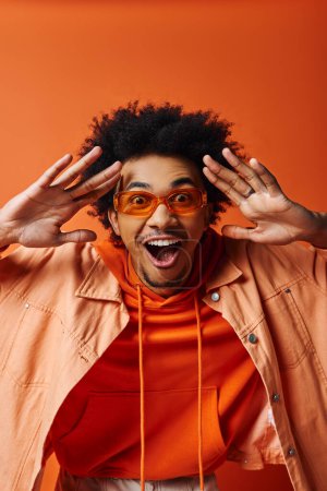 Stilvoller afroamerikanischer Mann mit lockigem Haar und trendiger Brille, der auf orangefarbenem Hintergrund einen witzigen Ausdruck macht.