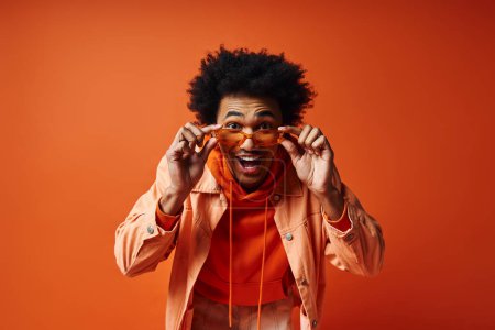 Photo for A stylish, young African American man in an orange jacket holds a pair of glasses against an orange background. - Royalty Free Image