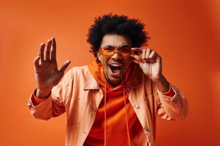 Ein stilvoller, junger afroamerikanischer Mann mit lockigem Haar und Sonnenbrille macht in einem orangefarbenen Hemd vor lebendigem Hintergrund eine lustige Miene..