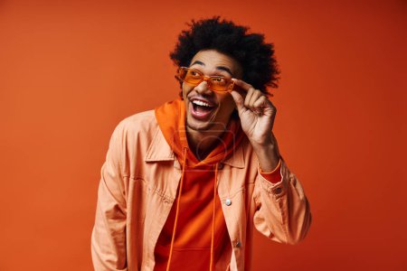 Téléchargez les photos : Un jeune Afro-Américain aux cheveux bouclés en tenue tendance et lunettes de soleil, mettant en valeur les émotions sur un fond orange. - en image libre de droit