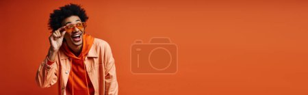 Ein stilvoller junger afroamerikanischer Mann mit lockigem Haar und modischer Kleidung verhüllt seine Augen mit einem orangefarbenen Schal vor orangefarbenem Hintergrund..