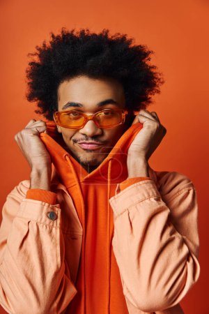Ein stilvoller junger afroamerikanischer Mann mit lockigem Haar trägt eine orangefarbene Jacke und eine trendige Sonnenbrille vor einem leuchtend orangen Hintergrund.