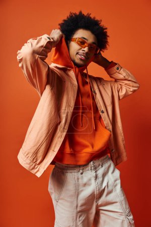 Foto de Un joven y elegante hombre afroamericano con el pelo rizado usando una camisa naranja y pantalones cortos blancos, exudando confianza sobre un fondo naranja. - Imagen libre de derechos