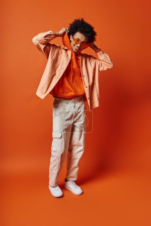 Foto de Hombre afroamericano rizado con camisa naranja de moda y pantalones caqui, estilo exuberante y carisma sobre un fondo naranja audaz. - Imagen libre de derechos