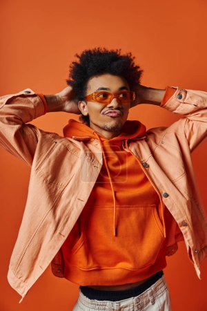 Foto de Un joven y elegante hombre afroamericano con un afro con una chaqueta y gafas de sol, exudando un ambiente fresco y seguro sobre un fondo naranja. - Imagen libre de derechos