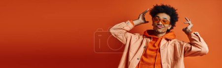 Ein stilvoller junger afroamerikanischer Mann mit lockigem Haar trägt orangefarbenes Hemd, Hose und Sonnenbrille und posiert mit emotionalem Ausdruck.