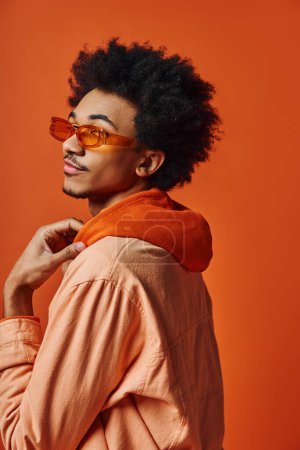 Ein stilvoller junger afroamerikanischer Mann mit lockigem Haar trägt Sonnenbrille und trendige Kleidung auf orangefarbenem Hintergrund.