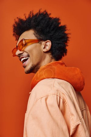 Ein stilvoller junger afroamerikanischer Mann mit lockigem Afro trägt Sonnenbrille und Jacke und drückt seine Emotionen auf orangefarbenem Hintergrund aus.