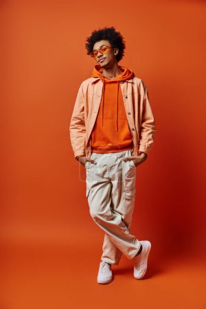 Trendy African American man with curly hair, wearing sunglasses, posing emotionally in bright orange hoodie and khaki pants.