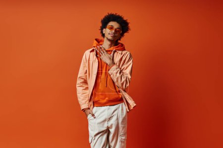 A trendy, curly African American man poses in an orange jacket and white pants against a bold orange backdrop, exuding confidence and style.