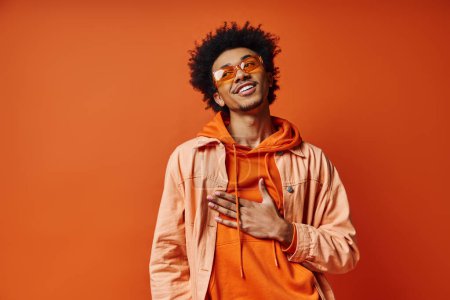 Photo for A curly African American man exudes emotion in trendy orange hoodie and sunglasses on bright orange backdrop. - Royalty Free Image