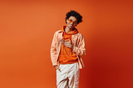 Stylish young African American man with curly hair wearing trendy orange jacket, white pants, and sunglasses on vibrant background.