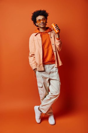 Téléchargez les photos : Un homme afro-américain branché avec des cheveux bouclés et des lunettes de soleil debout en toute confiance sur un fond orange vif. - en image libre de droit
