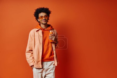 Ein angesagter afroamerikanischer Mann in orangefarbenem Kapuzenpullover und Sonnenbrille genießt einen Drink vor einem leuchtend orangen Hintergrund.