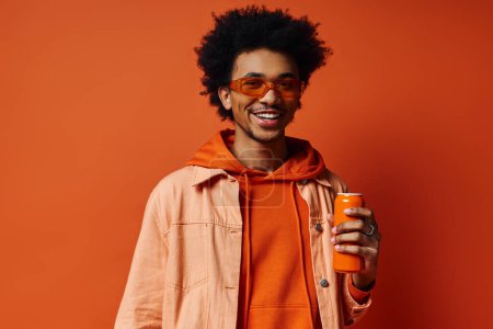 Homme afro-américain à la mode en sweat à capuche orange tenant une boîte de jus d'orange sur fond orange vif.