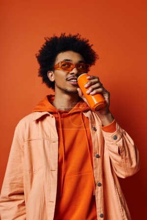 Photo for A trendy African American man, curly hair, orange hoodie, drinks from a can, sunglasses, emotional expression on orange background. - Royalty Free Image