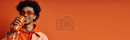 Foto de Elegante joven afroamericano en sudadera con capucha naranja de moda sosteniendo una lata de refresco contra un fondo naranja vibrante. - Imagen libre de derechos