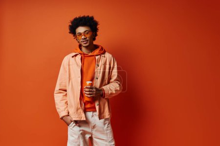 Téléchargez les photos : Jeune homme afro-américain élégant posant avec confiance dans une veste orange à la mode et un pantalon blanc sur un fond orange vif. - en image libre de droit