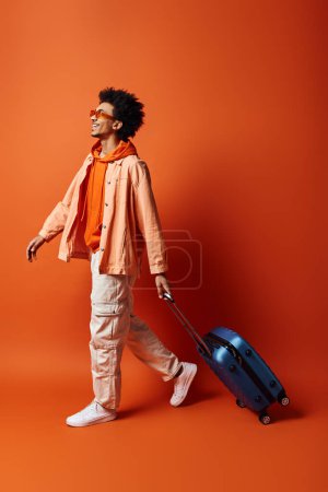A stylish young African American man with curly hair and trendy attire walking forward while holding a suitcase in his hand against an orange background.