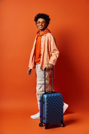 A stylish young African American man standing with a suitcase in front of an orange wall, exuding confidence and charisma.