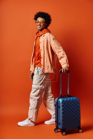 Foto de Un hombre afroamericano de moda con una maleta se levanta con confianza contra un vibrante telón de fondo naranja, exudando un sentido de aventura y estilo. - Imagen libre de derechos