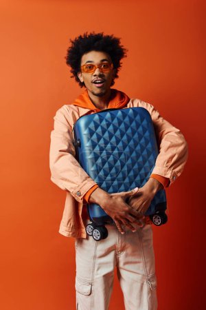 Stylish African American man in trendy attire and sunglasses holding a blue piece of luggage on an orange background.