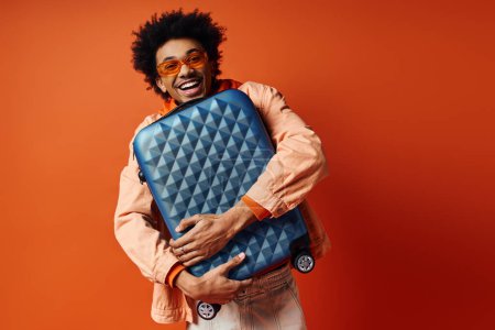 A stylish young African American man with curly hair and trendy attire stands against an orange background, holding a blue piece of luggage.