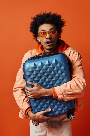 A stylish young African American man with curly hair holding a blue piece of luggage against an orange background.