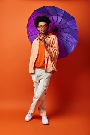 Ein junger afroamerikanischer Mann mit lockigem Haar hält einen lila Regenschirm vor einem kühnen orangefarbenen Hintergrund, der Stil und Persönlichkeit ausstrahlt.