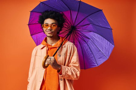 A curly African American man in trendy attire and sunglasses, holding a purple umbrella on an orange background.
