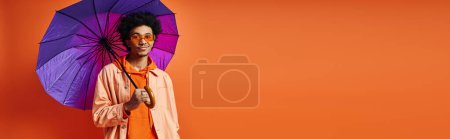 Foto de Un joven afroamericano de moda con pelo rizado, gafas de sol y atuendo de moda sostiene un vibrante paraguas púrpura sobre un fondo naranja. - Imagen libre de derechos