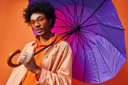 Joven afroamericano con el pelo rizado y gafas de sol sosteniendo un paraguas púrpura sobre su cabeza sobre un fondo naranja.