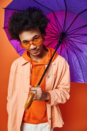 Foto de Un joven afroamericano de moda con el pelo rizado sosteniendo un paraguas púrpura, exudando emoción sobre un fondo naranja. - Imagen libre de derechos