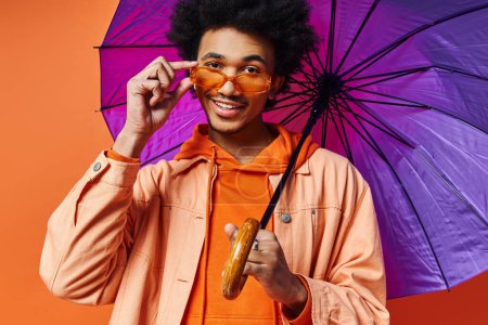 A trendy young African American man with curly hair, wearing sunglasses, holding an umbrella, and smiling at the camera on an orange background.