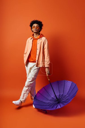 Foto de Un hombre afroamericano joven y elegante con el pelo rizado está junto a un paraguas púrpura sobre un fondo naranja. - Imagen libre de derechos