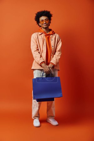 Curly, trendy African American man holding a blue bag in front of orange backdrop, showcasing emotions and style.