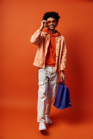 Foto de Joven hombre afroamericano con atuendo moderno, pelo rizado y gafas de sol, sosteniendo una bolsa azul mientras habla en un teléfono celular. - Imagen libre de derechos