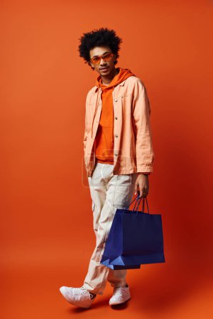 Ein junger Afroamerikaner in leuchtend orangefarbenem Hemd und weißer Hose hält eine blaue Tasche vor leuchtend orangefarbenem Hintergrund.