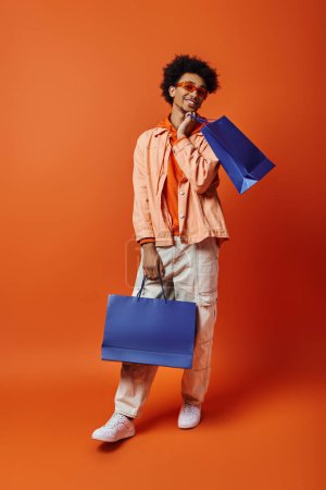 Lockiger afroamerikanischer Mann in trendiger Kleidung hält eine blaue Tasche in der Hand, während er eine einzigartige, lebendige blaue Tasche vor orangefarbenem Hintergrund präsentiert.